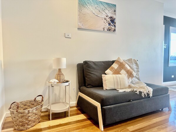 Ein modernes Wohnzimmer mit einem grauen Sofa, einem kleinen Beistelltisch mit Lampe, einem Korbwäschesammler und einem gerahmten Bild an der Wand.