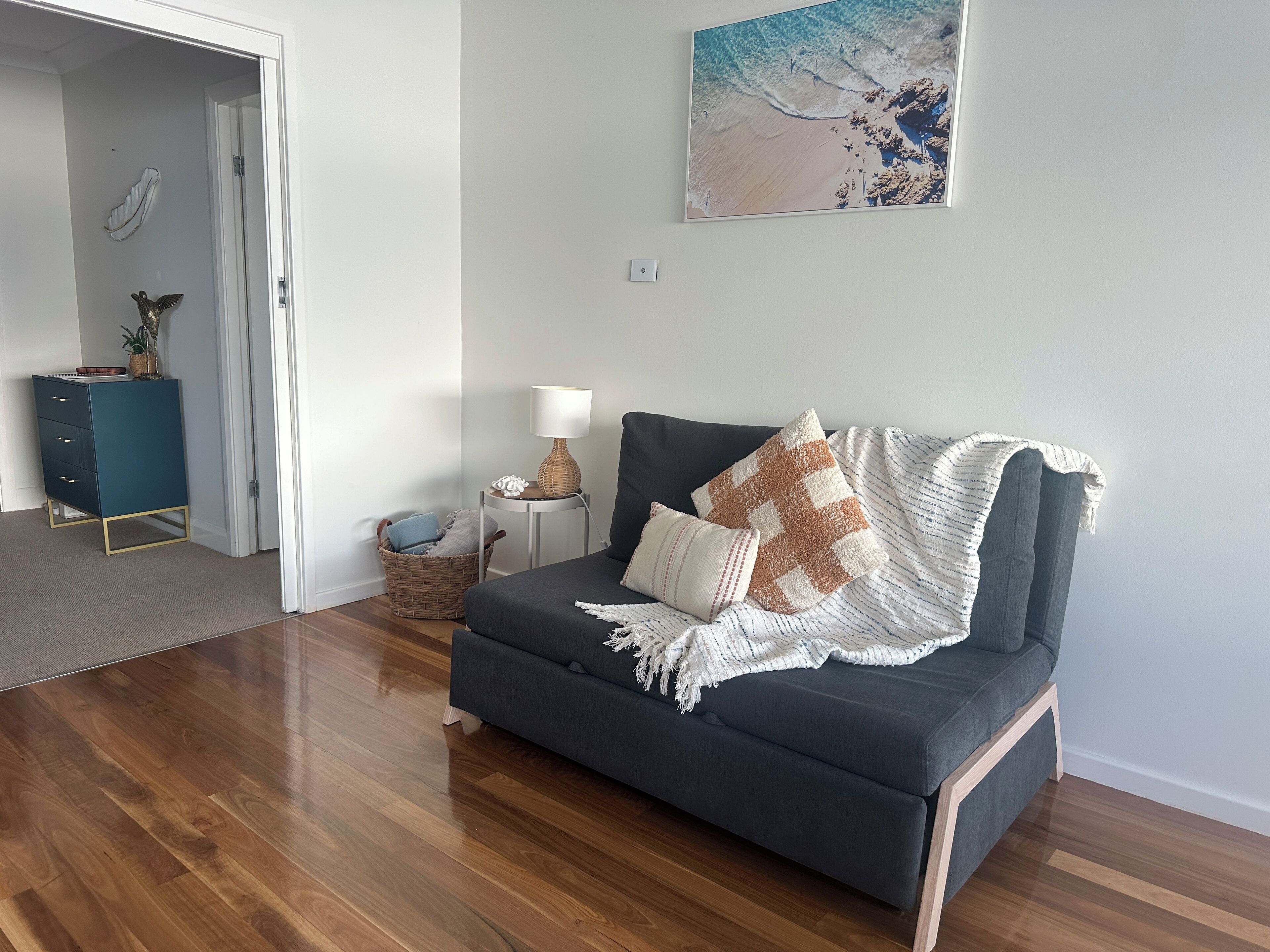 A modern living room with a grey sofa, a small side table with a lamp, a wicker basket, and a wall-mounted picture of a beach.