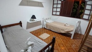 Un petit logement composé d’une seule pièce, avec un lit en bois, une table de chevet avec une lampe, une petite fenêtre avec des volets et un sol à motifs.