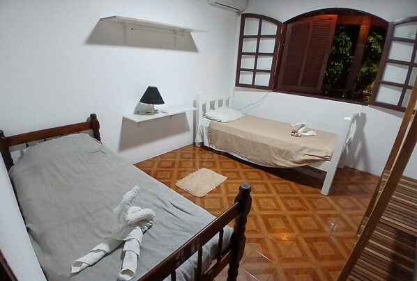 A small, single-room accommodation with a wooden bed, a bedside table with a lamp, a small window with shutters, and a patterned floor.