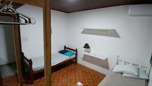 Un petit logement composé d’une seule pièce, avec un lit superposé en bois, une table de chevet avec une lampe, une étagère au-dessus du lit et un appareil de climatisation blanc fixé au mur.