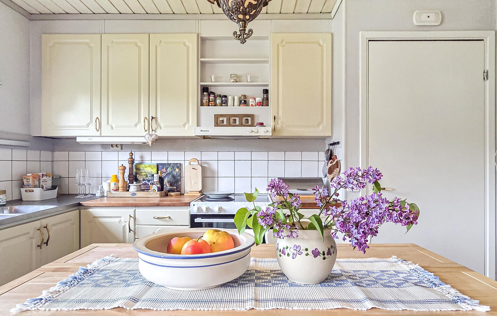 Ett kök med vita skåp, en spis och ett diskho. På köksbänken står en skål med frukt och en vas med lila blommor.