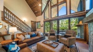 A spacious living room with a high ceiling, wooden staircase, and large windows offering a view of the outdoors.