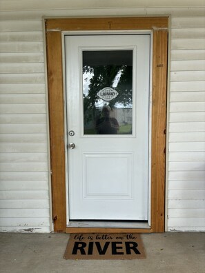 A white door with a wooden frame, a window with a laundry sign, and a doormat with the text 'life is better on the river'.