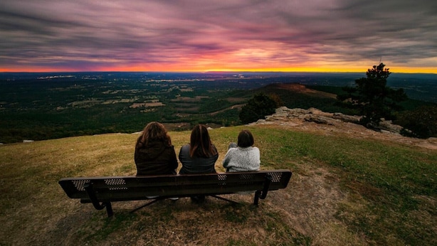 Trois personnes sont assises sur un banc, face à un paysage illuminé par le coucher du soleil.