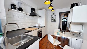 Una cocina moderna con un lavamanos, estufa y encimera negra. Hay armarios blancos, una campana extractora negra y una zona de comedor blanca con una silla.