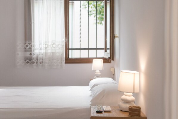 A neatly made bed with white linens, a bedside lamp, and a remote control on a wooden nightstand.