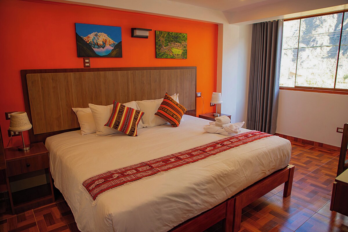 Una camera d'albergo con un letto grande, una testiera in legno, due quadri appesi e un comodino con una lampada, oltre a una finestra con tende.