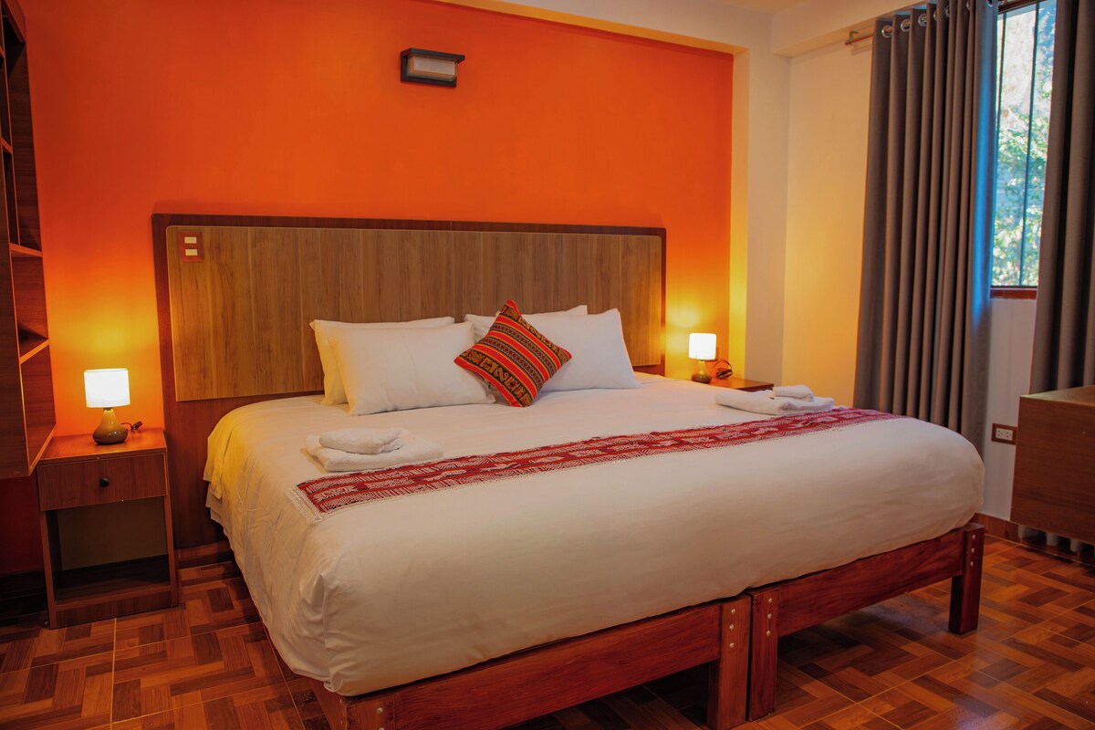 A hotel room with a large bed, wooden headboard, two bedside tables with lamps, a red patterned throw, and a view of the outdoors through a window with curtains.