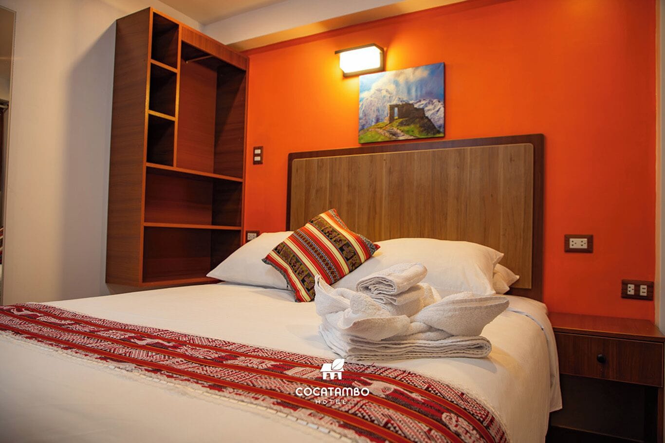 A hotel room with a large bed, a wooden headboard, a colorful pillow, and a painting on the wall.