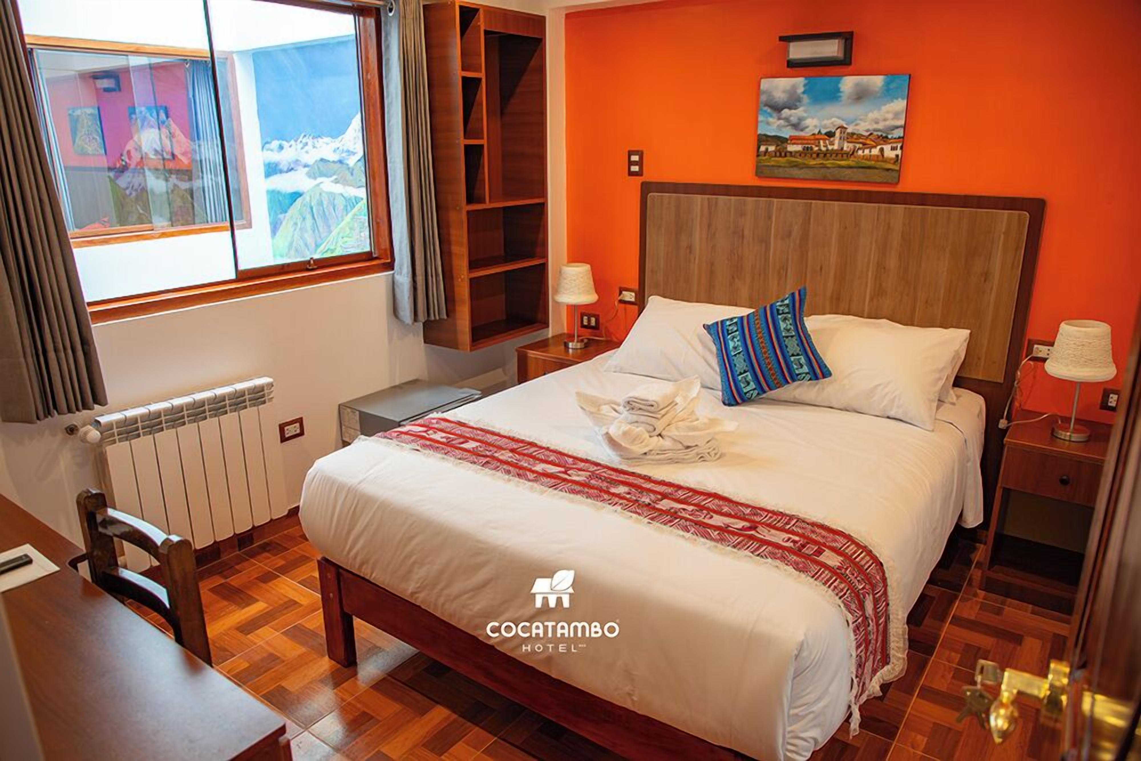 A hotel room with a wooden bed, a window with a view of mountains, a wooden nightstand, and a red wall.