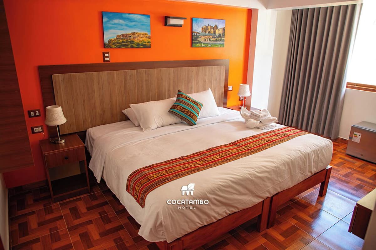 A hotel room with a wooden bed, white bedding, a colorful throw blanket, and two paintings on the orange wall.