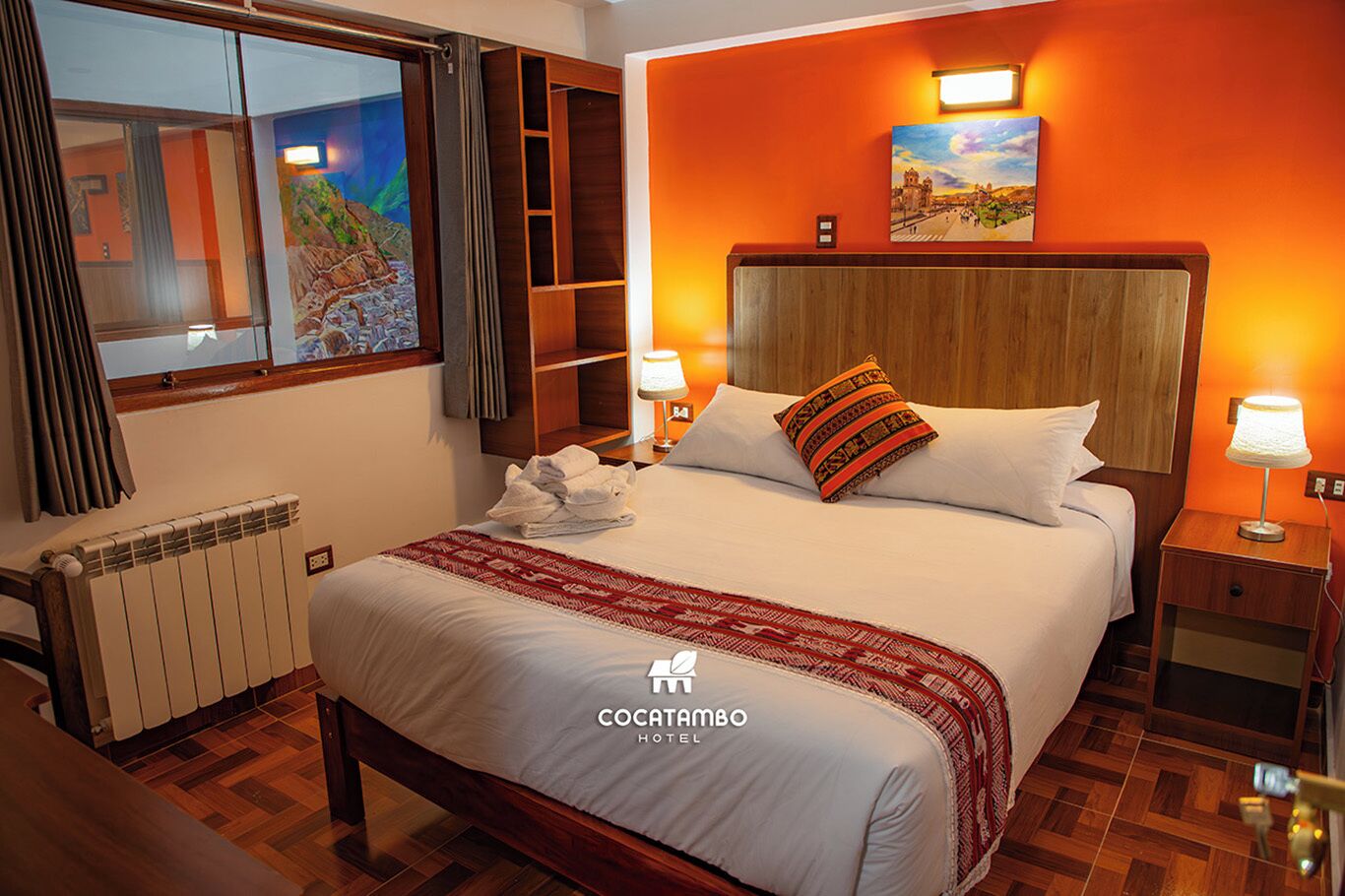 A hotel room with a large bed, two bedside lamps, a painting above the bed, a radiator, and a window with curtains.