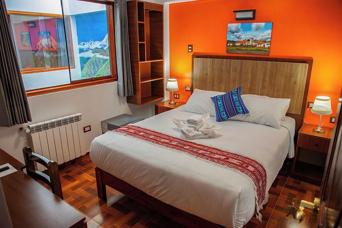 A hotel room with a bed, bedside lamps, a view of mountains through a window, and a wooden headboard.