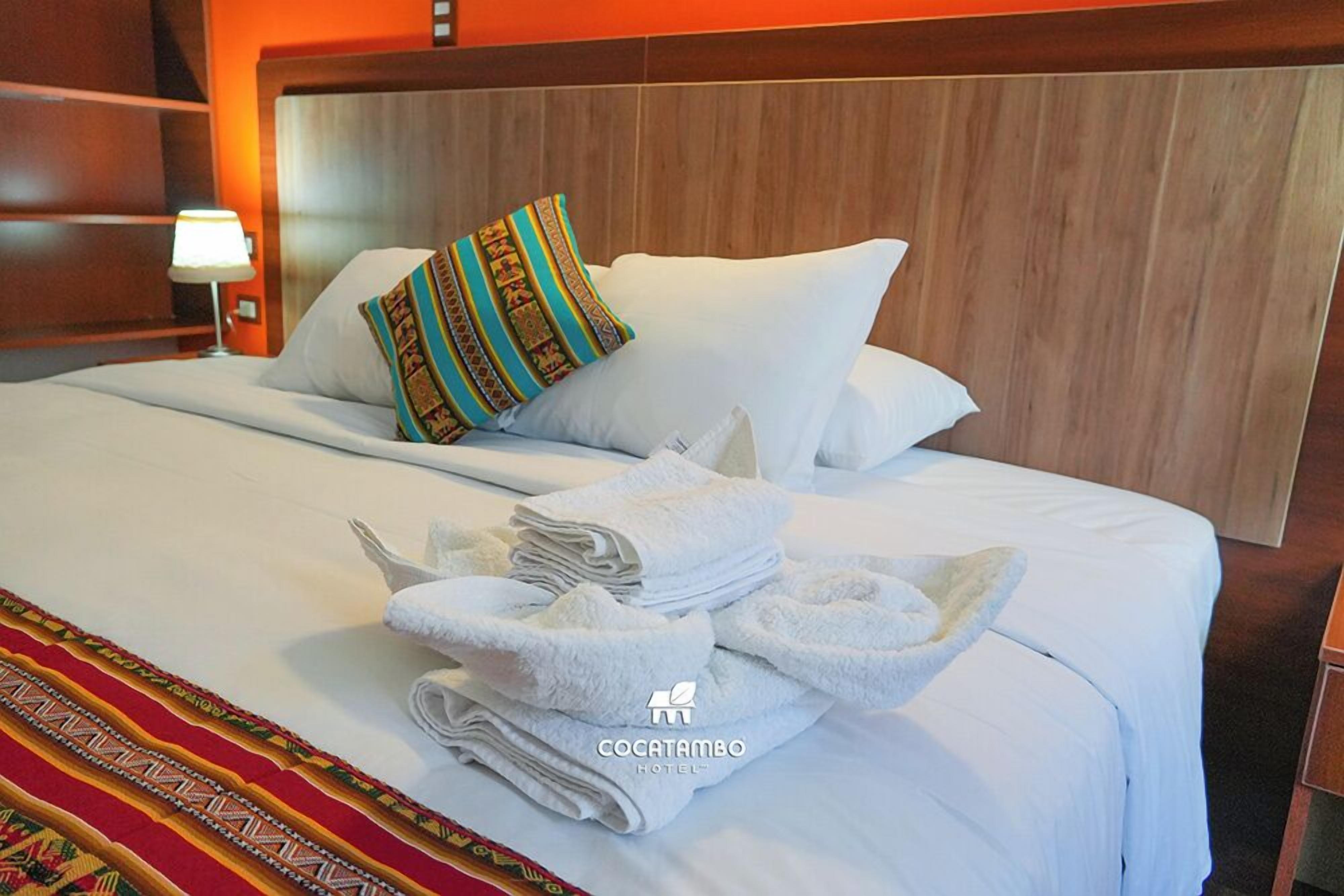 A hotel room with a neatly made bed, a colorful pillow, and a stack of towels.