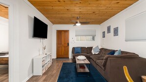 A modern living room with a wooden ceiling, a flat-screen TV, a brown sofa, a wooden coffee table, and a blue rug. - Midtown Reno 1br W/ share Hot Tub & Backyard (Reno)
