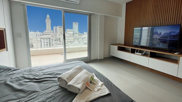 A modern hotel room with a large bed, a flat-screen TV, a sliding glass door to a balcony, and a city view. - Studio Luxury Balcony Pool (Buenos Aires)