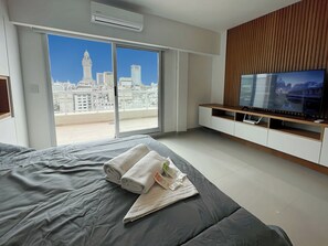 A modern hotel room with a large bed, a flat-screen TV, a sliding glass door to a balcony, and a city view. - Studio Luxury Balcony Pool (Buenos Aires)