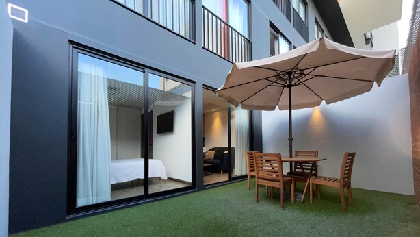 A modern hotel room with a sliding glass door leading to a balcony with artificial grass, a wooden table with chairs, and a large umbrella providing shade.