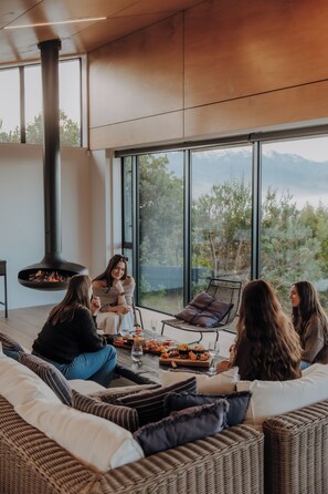 A modern interior with a large window, a fireplace, wicker furniture, and a dining table with food and drinks.