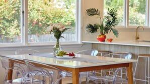 A dining area with a wooden table and clear chairs, a vase with flowers, and a view of greenery through large windows. - Close To Beach | Sunny | Getaway - KK16786 (Kaikoura)