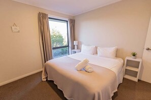 Ein Hotelzimmer mit einem großen Bett, einem Fenster mit Vorhängen, einem Nachttisch mit Lampe und einer kleinen Pflanze auf einem Beistelltisch.