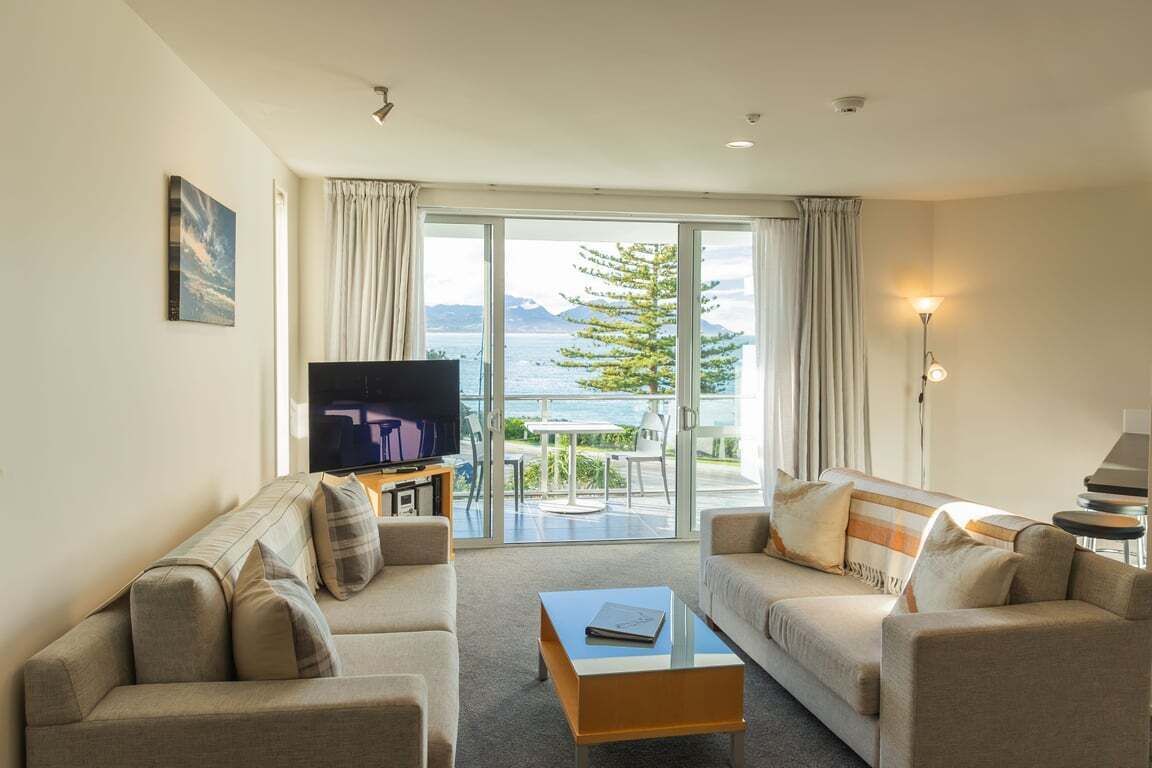 Un salon moderne comprenant un ensemble de canapés, une table basse, un téléviseur à écran plat et une vue sur la mer grâce à de grandes fenêtres.