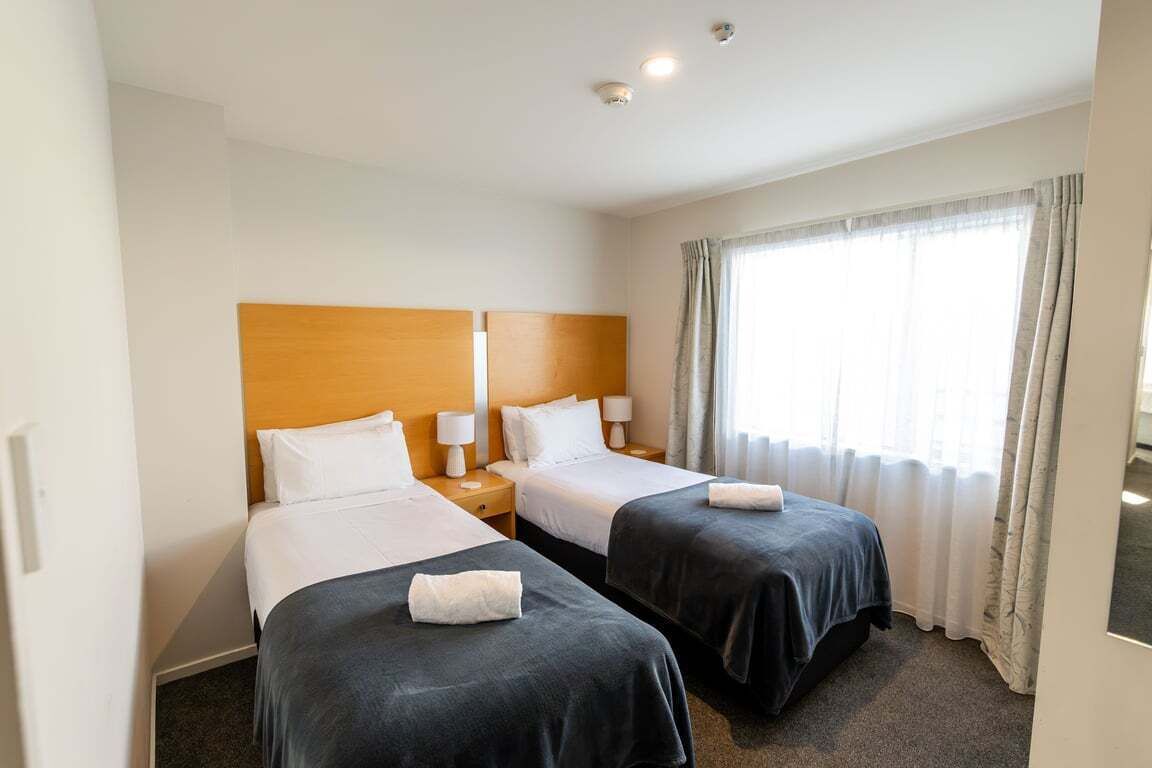 Een hotelkamer met twee bedden, een houten hoofdbord, bedlampen en een groot raam met gordijnen.