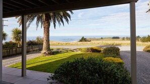 A covered outdoor area with a view of a grassy field, a palm tree, and a distant body of water. - The Lookout | Spacious | Stunning Views - KK12632 (Kaikoura)