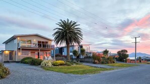 A modern two-story house with a large balcony, surrounded by a well-maintained lawn and palm trees. - The Lookout | Spacious | Stunning Views - KK12632 (Kaikoura)
