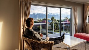 A person seated in a wicker chair by a large window overlooking a mountain landscape. - The Lookout | Spacious | Stunning Views - KK12632 (Kaikoura)