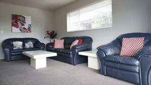 Ein Wohnzimmer mit zwei schwarzen Ledersofas, weißen Couchtischen und roten und weißen Kissen. An der Wand hängt ein farbenfrohes abstraktes Gemälde, und es gibt ein Fenster mit Jalousien.