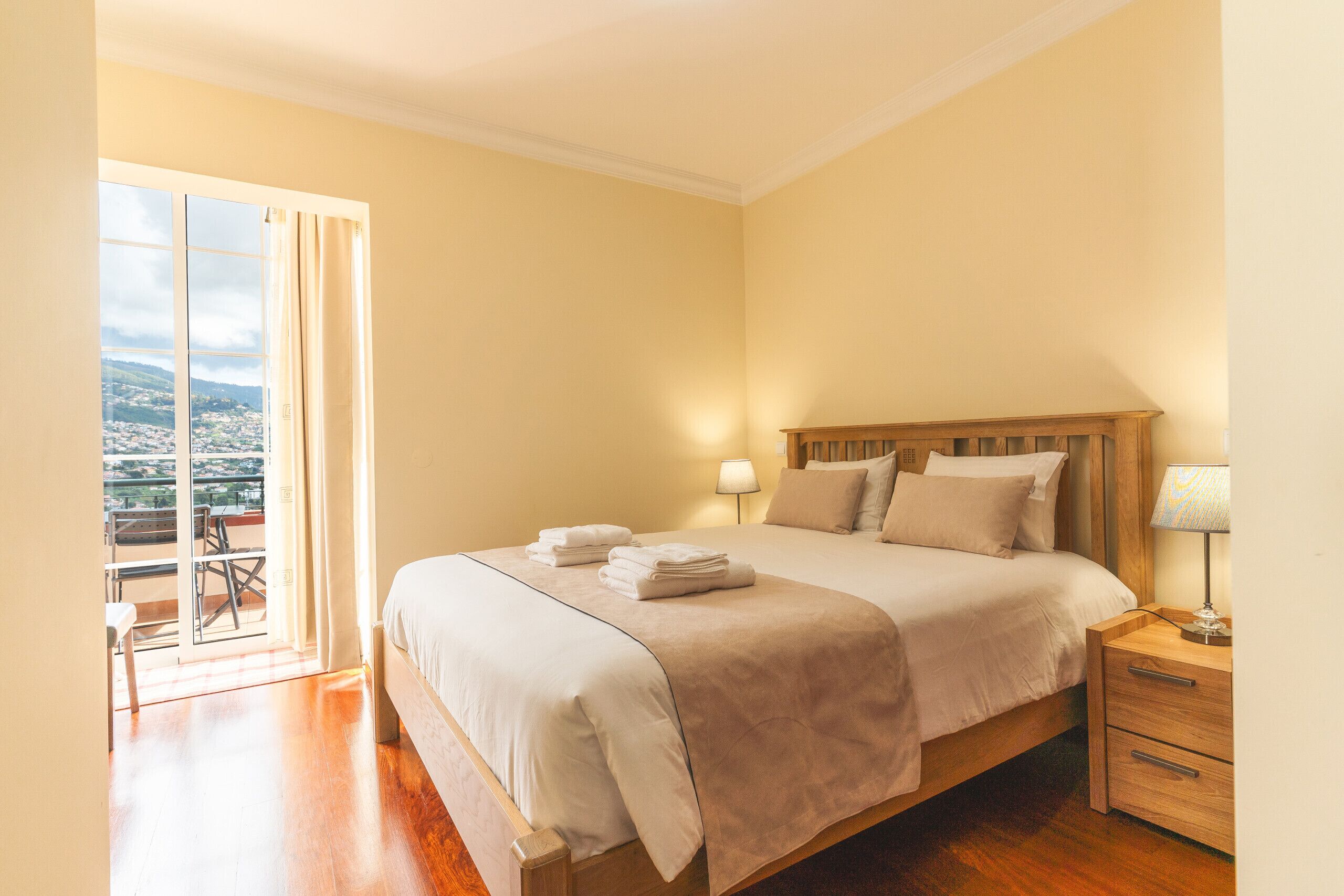 A neatly made bed with a wooden headboard, a bedside table with a lamp, and a view of a balcony with outdoor furniture.