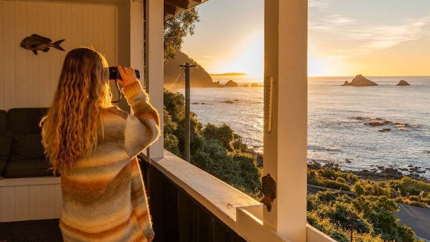 Una persona fotografiando una vista costera desde un balcón con barandilla de madera y un sofá.
