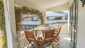 Een overdekt terras met een tafel en stoelen, omgeven door een witte hek en begroeiing.
