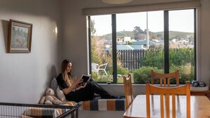A person sitting on a bench by a window with a view of a fenced garden and hills. - Handy to Town | Spacious | Relaxed - KK14905 (Kaikoura)