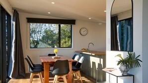Uma sala de jantar moderna com mesa e cadeiras de madeira, uma janela grande com vista para árvores, um relógio na parede e uma pequena planta sobre uma mesinha.