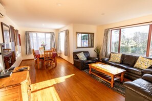 A spacious living room with a wooden floor, a sofa set, a dining table, and a television. - Coastal | Spacious | Mountain Views - KK21238 (Kaikoura)