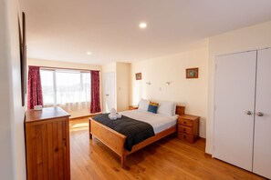 A bedroom with a wooden bed, a wardrobe, a bedside table, and a window with curtains. - Coastal | Spacious | Mountain Views - KK21238 (Kaikoura)