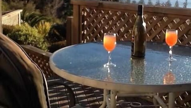 A round glass table with two glasses of orange beverage and a bottle, set against a backdrop of a mountainous landscape and a body of water. - Spectacular Views | Convenient | Affordable - KK17129 (Kaikoura)