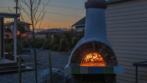 A brick outdoor oven with a fire burning inside, surrounded by a stone base and a metal grill. - Kowhai Cottage | Pizza Oven | Super Central - KK32726 (Kaikoura)