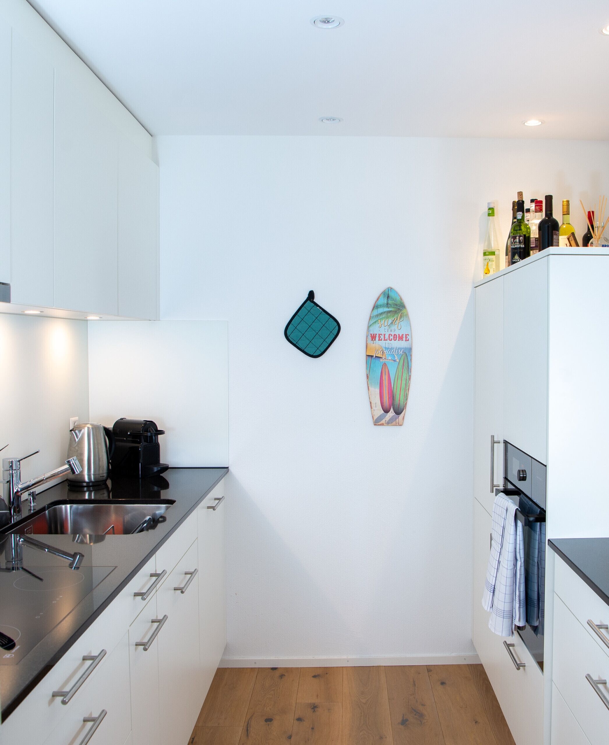 A modern kitchen with white cabinets, a stainless steel sink, and a coffee machine. There is a surfboard decoration on the wall and a rack of bottles on the countertop.