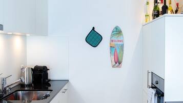 A modern kitchen with white cabinets, a stainless steel sink, and a coffee machine. There is a surfboard decoration on the wall and a rack of bottles on the countertop.