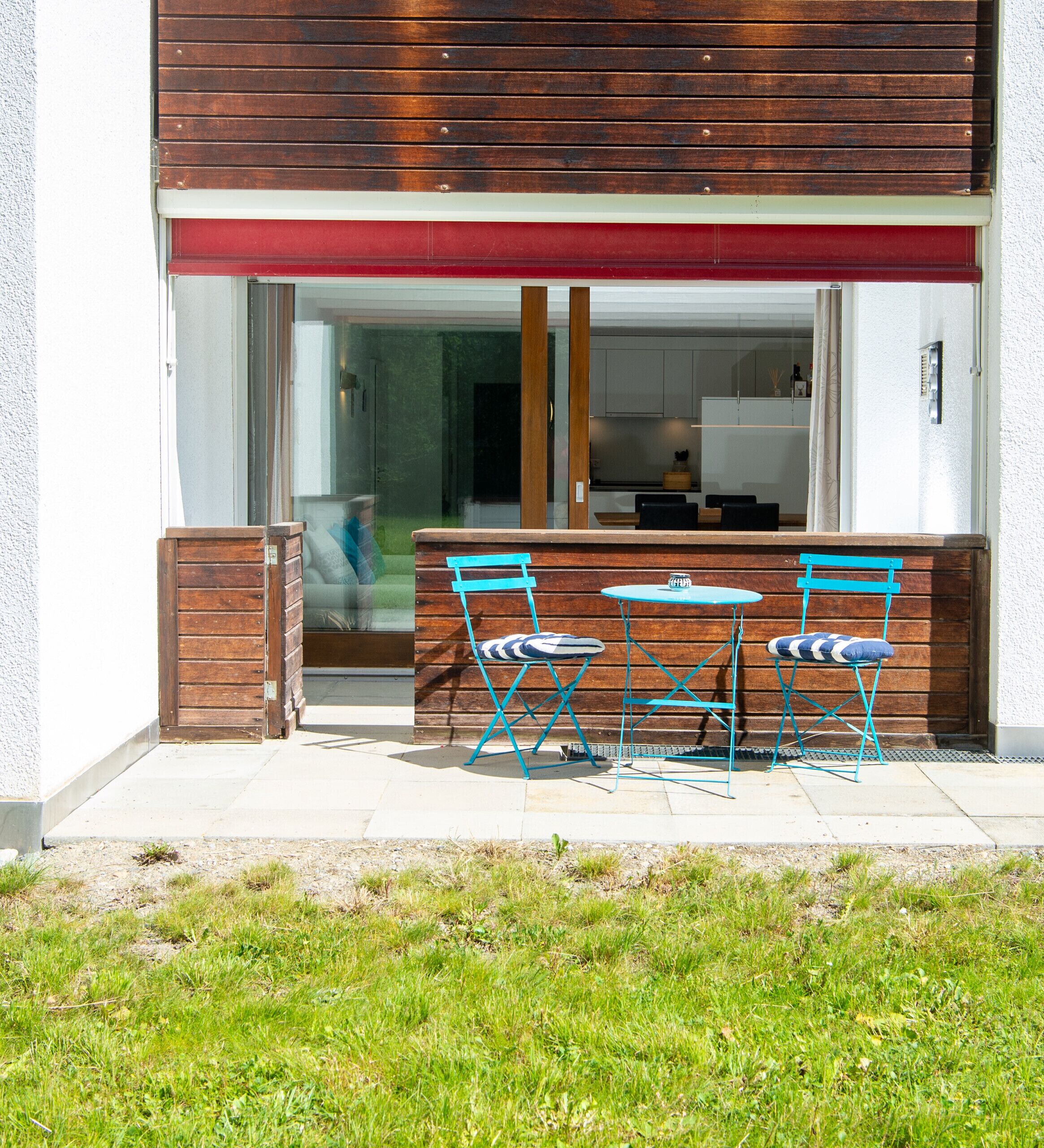 Ein modernes Gebäude mit Holzfassade, rotem Markisenvorbau und Glastür, die in einen Küchenbereich führt. Vor der Tür stehen zwei blaue Klappstühle und ein kleiner runder Tisch.