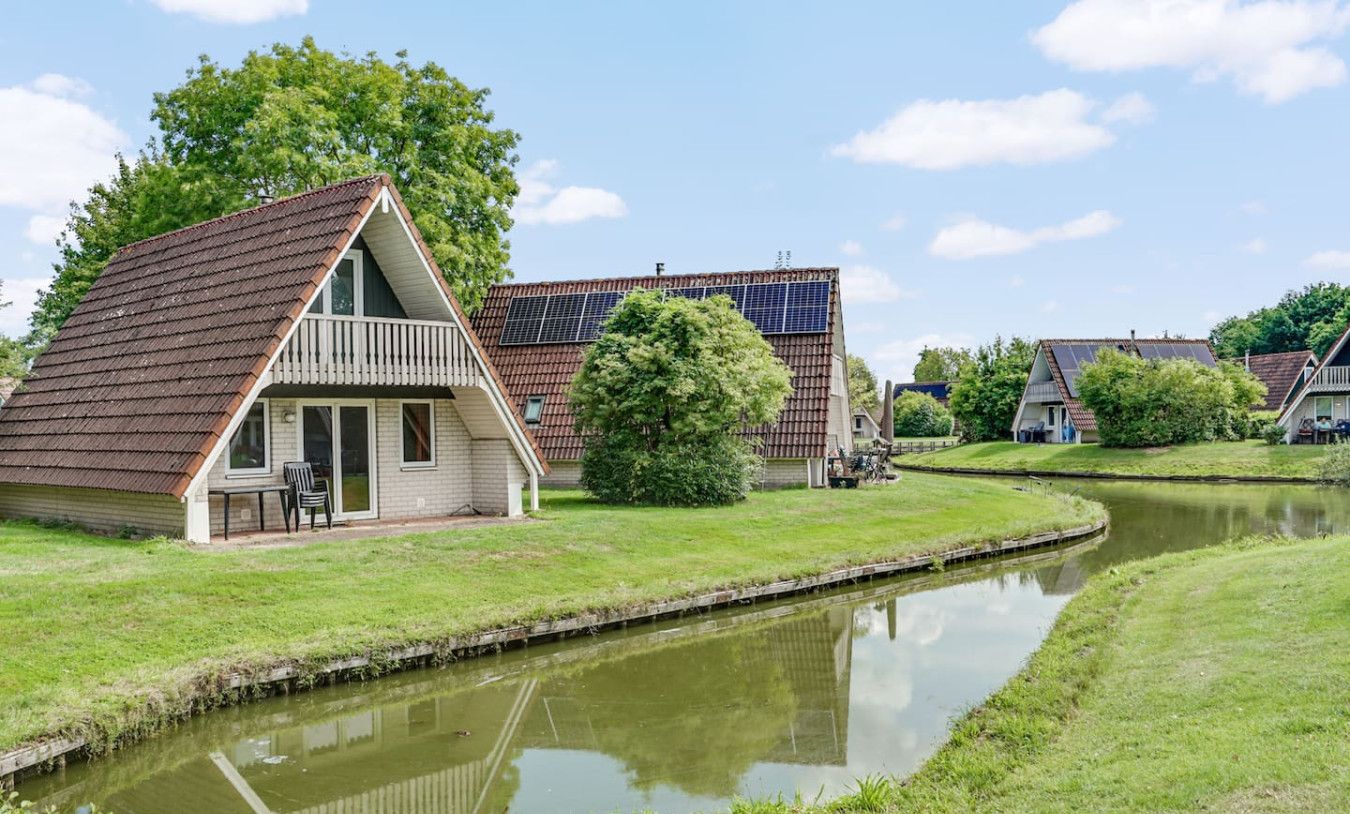 Un edificio junto a un canal, con un balcón, al lado de una casa con paneles solares, y una masa de agua tranquila que refleja el entorno.