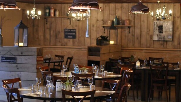 Un restaurant aux murs en bois et aux lustres étincelants, où les tables sont dressées avec des verres à vin et des assiettes, et où un panneau annonce un menu spécial.
