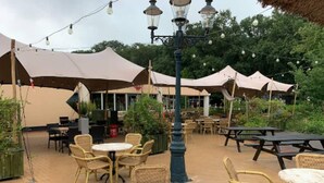 En udendørs terrasse med runde borde og fletstole, omgivet af store parasoller og lyskæder.