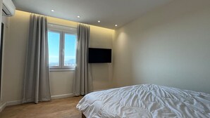 Ein Schlafzimmer mit einem Bett, einem Fenster mit Vorhängen, einem an der Wand befestigten Fernseher und einer Klimaanlage.