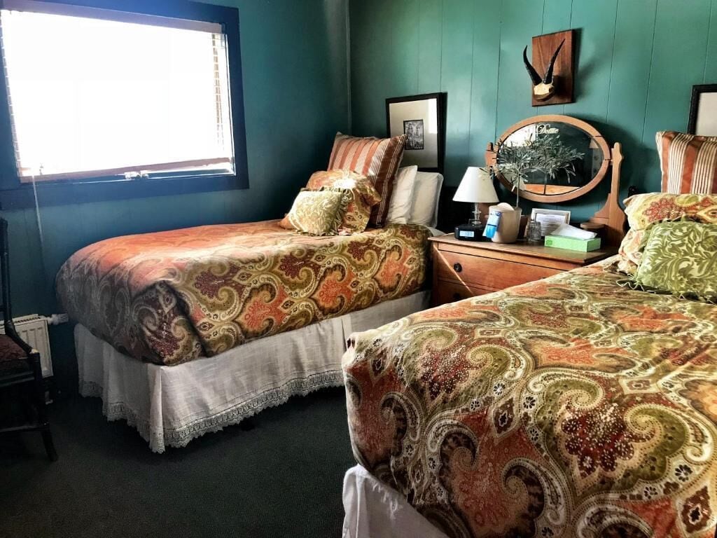A room with two beds, a window, a bedside table with a lamp, a mirror, and a framed picture on the wall.