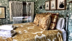 Ein Schlafzimmer mit blumigem Tapetenmuster, einem Bett mit geblümter Bettwäsche, einem Fenster mit transparenten Vorhängen und gerahmten Bildern an der Wand.